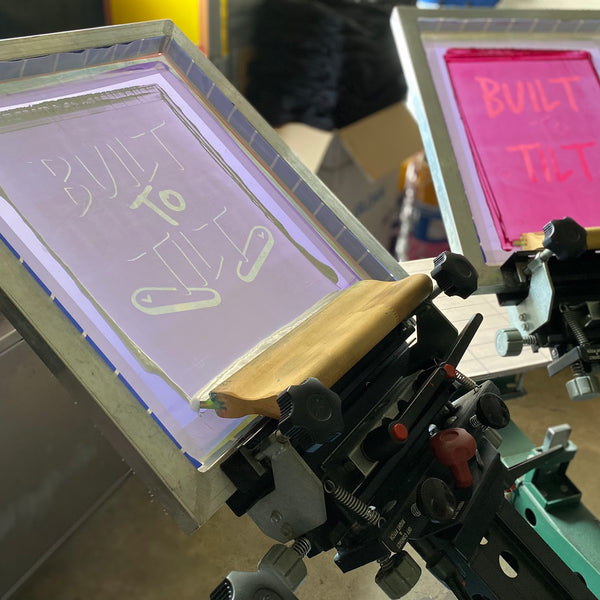 Built to Tilt screens on the press, one coated in pink ink with a squeegee on the white screen at RVA Threads screen printing, Virginia
