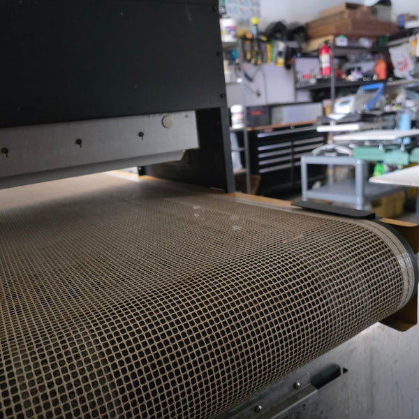 Mesh belt of the conveyor dryer running into the heat chamber with the press in the background at RVA Threads screen printing, Virginia