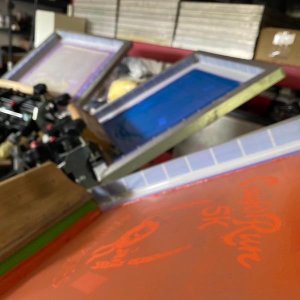 Crush Run orange ink screen with the squeegee resting on the press at RVA Threads screen printing, Virginia