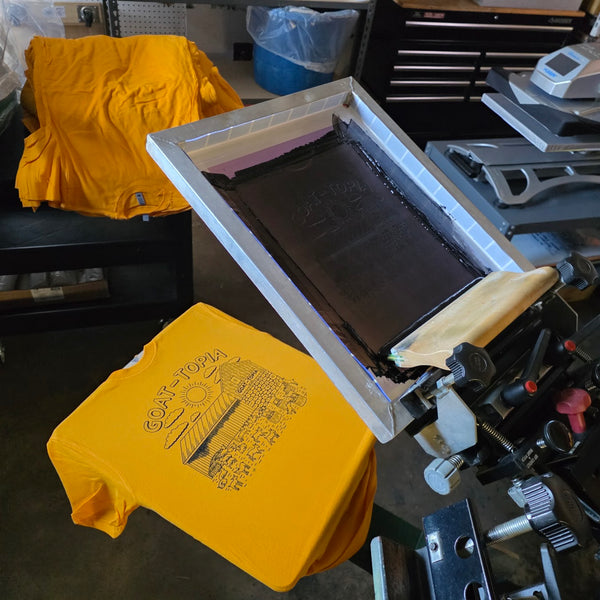 Goat-Topia print in black ink on a gold Next Level 3600 tee, on the press with the screen raised at RVA Threads screen printing, Virginia