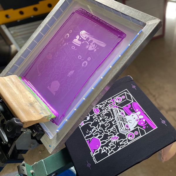 Meow Royale DJ bunny screen raised over a printed black tee with the squeegee on the press at RVA Threads screen printing, Virginia