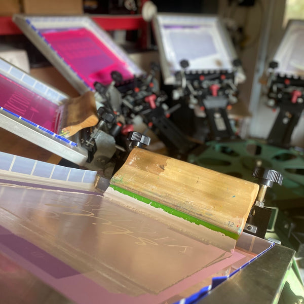 Pink ink screens loaded on the press with a wooden squeegee resting on the front screen at RVA Threads screen printing, Virginia