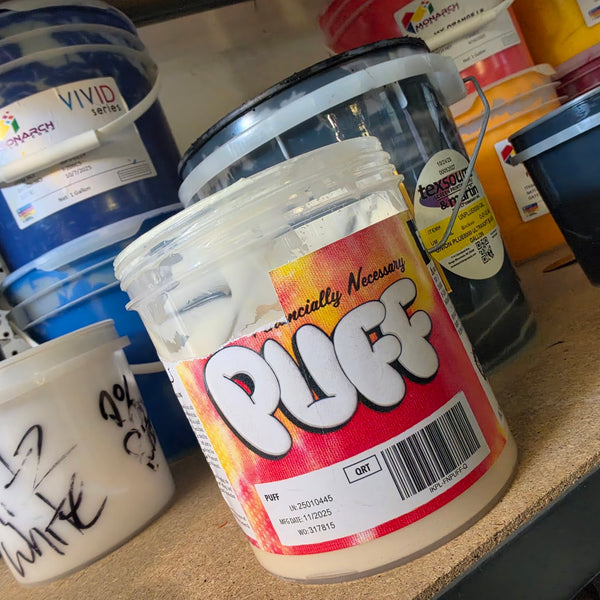 Tub of puff ink in front of a shelf of plastisol ink buckets in blue, orange, red and white at RVA Threads screen printing, Virginia