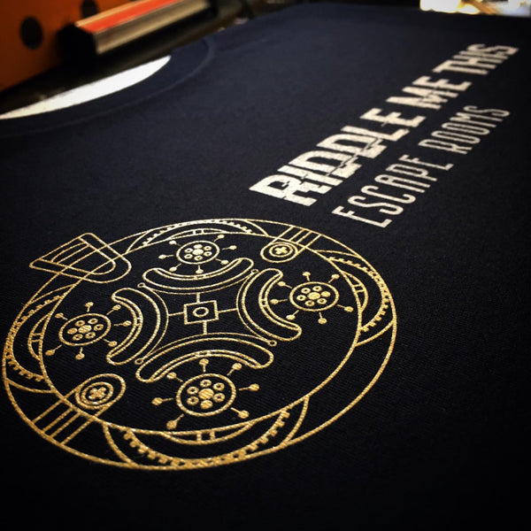 Riddle Me This Escape Rooms logo in gold ink on a navy tee, laid flat at RVA Threads screen printing, Virginia
