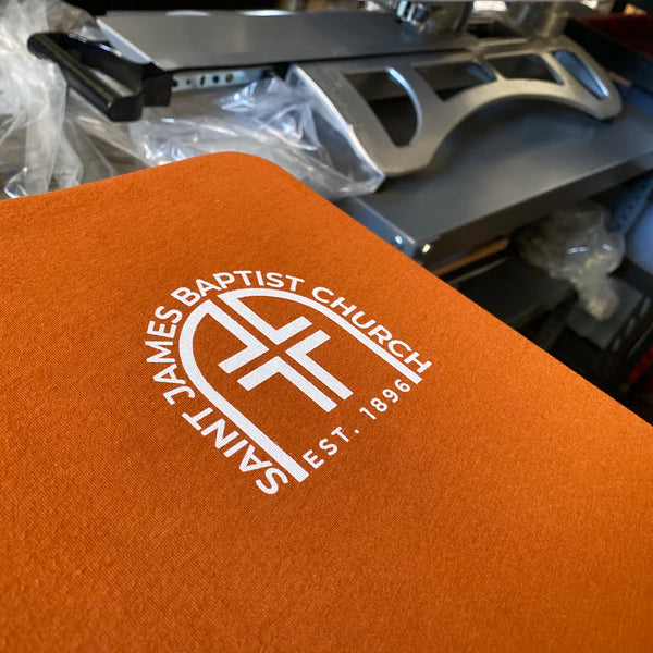 Saint James Baptist Church logo in white ink on an orange tee at RVA Threads screen printing, Virginia