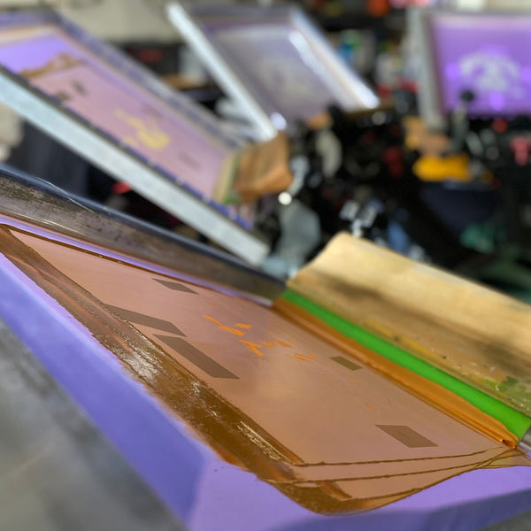 Screen flooded with gold ink and the squeegee resting on the press between prints at RVA Threads screen printing, Virginia