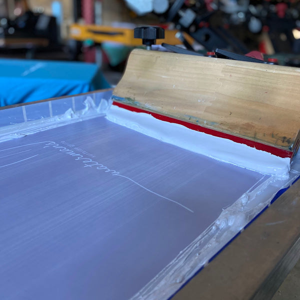 Screen with white ink and the squeegee over a blue tee on the press at RVA Threads screen printing, Virginia