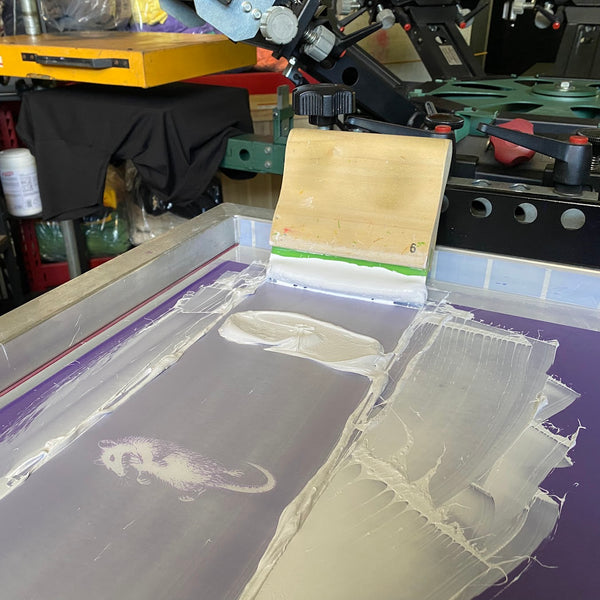 Screen with white ink and the squeegee resting on the press between prints at RVA Threads screen printing, Virginia