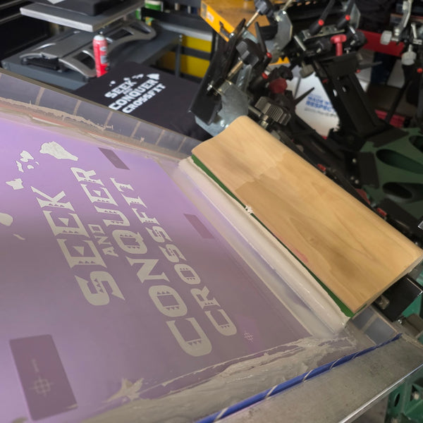 Seek and Conquer CrossFit screen with a wood squeegee on the press, a printed Black hoodie behind at RVA Threads screen printing, Virginia