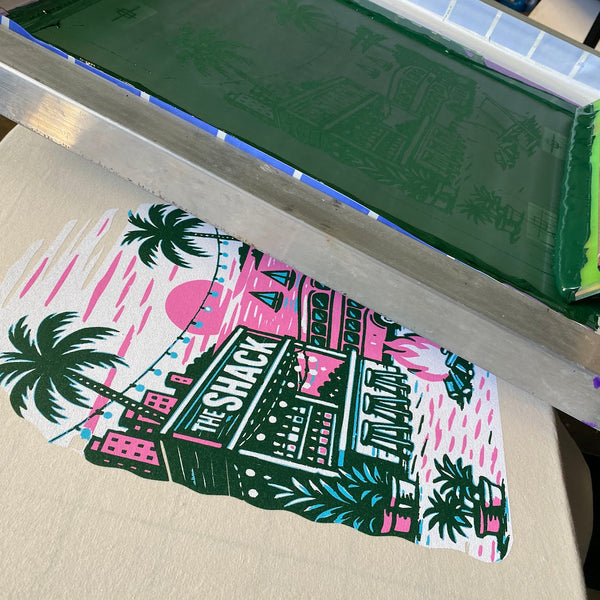 The Shack screen with green ink pulled over a printed tee on the press at RVA Threads screen printing, Virginia