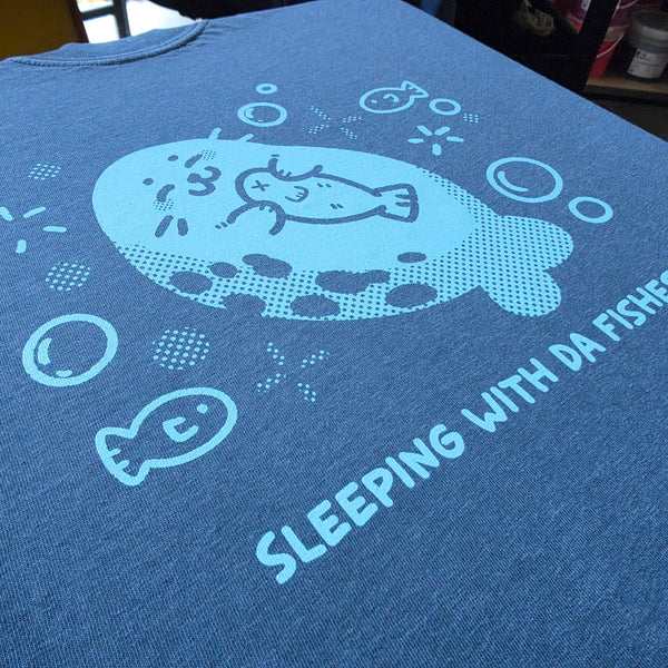 Close up of the Sleeping With Da Fishes seal print in light blue on a Denim Comfort Colors 1717 tee at RVA Threads screen printing, Virginia