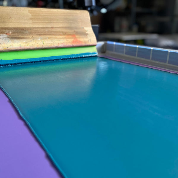 Teal ink flooded across a screen with the squeegee on the press at RVA Threads screen printing, Virginia
