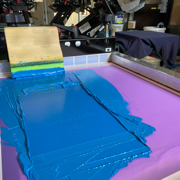 Blue ink spread across a screen with the squeegee on the press at RVA Threads screen printing, Virginia