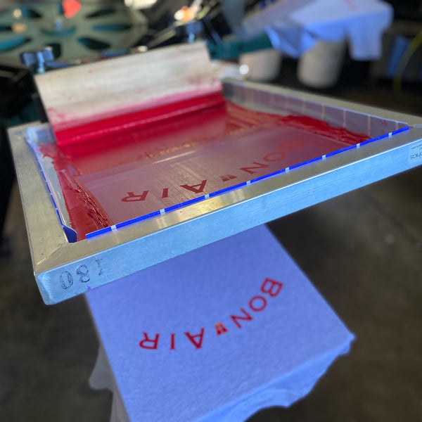 Bon Air screen with red ink and squeegee on the press over a printed tee at RVA Threads screen printing, Virginia