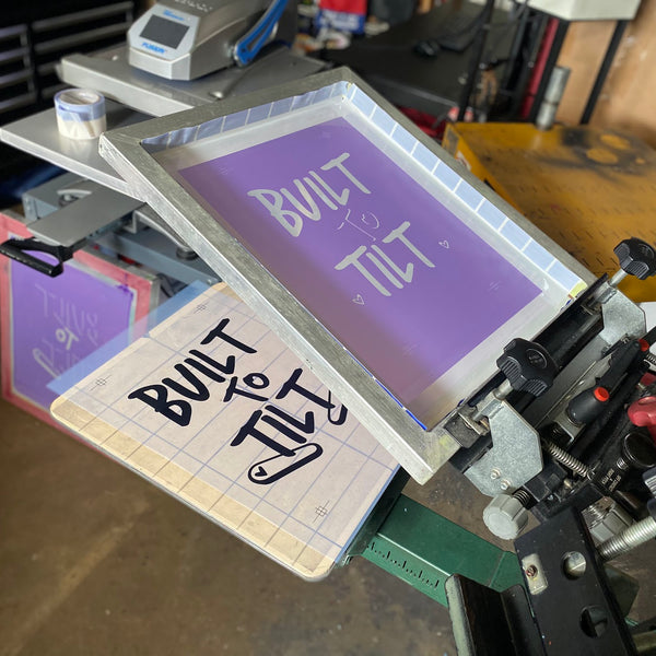 Built to Tilt screen raised on the press beside a printed proof of the design at RVA Threads screen printing, Virginia