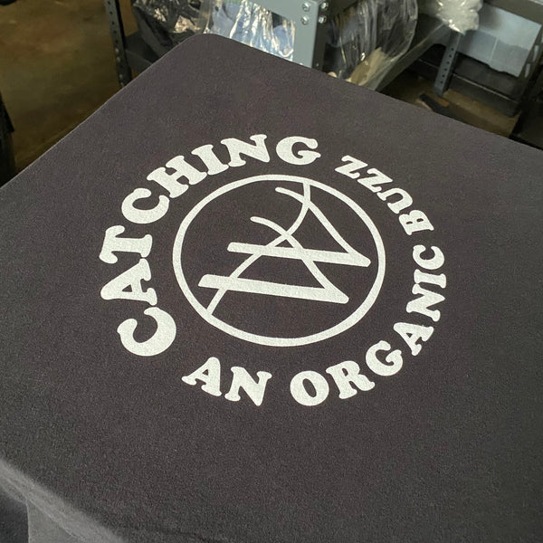 Catching An Organic Buzz logo in white ink on a charcoal tee on the press pallet at RVA Threads screen printing, Virginia