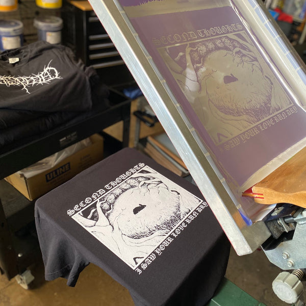 Circular eye graphic in white ink on black tees with the screen raised on the press at RVA Threads screen printing, Virginia