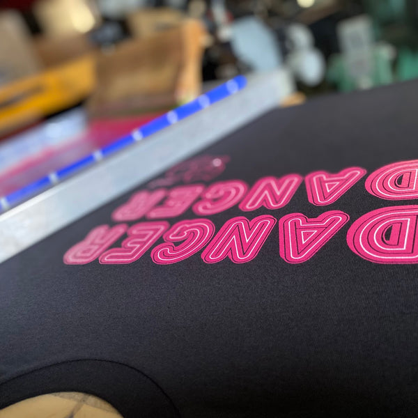 Danger Danger design in neon pink ink on a black Bella+Canvas 3001 tee at the press at RVA Threads screen printing, Virginia