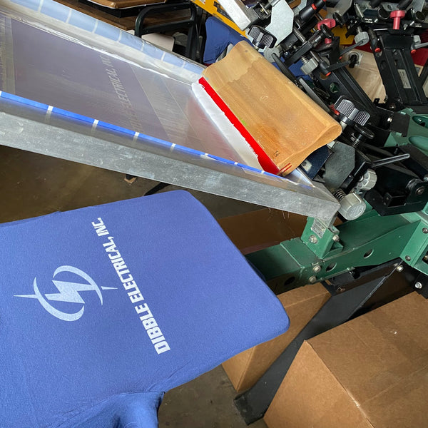 Dibble Electrical, Inc. logo in white ink on a blue tee under the screen and squeegee on the press at RVA Threads screen printing, Virginia