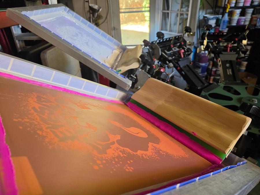 Eyes Peeled pink ink screen and wood squeegee on the press, white underbase screen raised behind at RVA Threads screen printing, Virginia