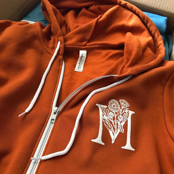 Hamilton Mercantile M monogram in white ink on an orange zip hoodie at RVA Threads screen printing, Virginia