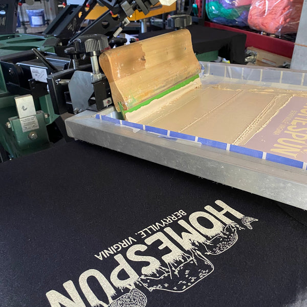 Homespun design in white ink on a black tee with the screen and squeegee raised above at RVA Threads screen printing, Virginia
