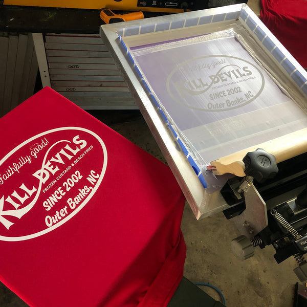 Kill Devils screen raised over a printed red tee on the press at RVA Threads screen printing, Virginia