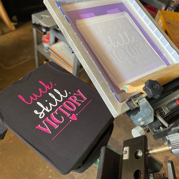 Luck Skill Victory design in pink and white ink on a black Bella+Canvas 3001 tee beside the press at RVA Threads screen printing, Virginia