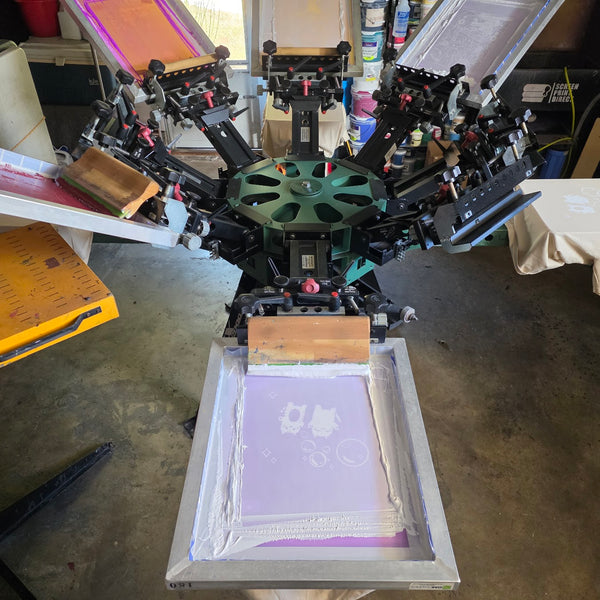 Manual press loaded with purple emulsion screens and wood squeegees, a cream tee on one platen at RVA Threads screen printing, Virginia