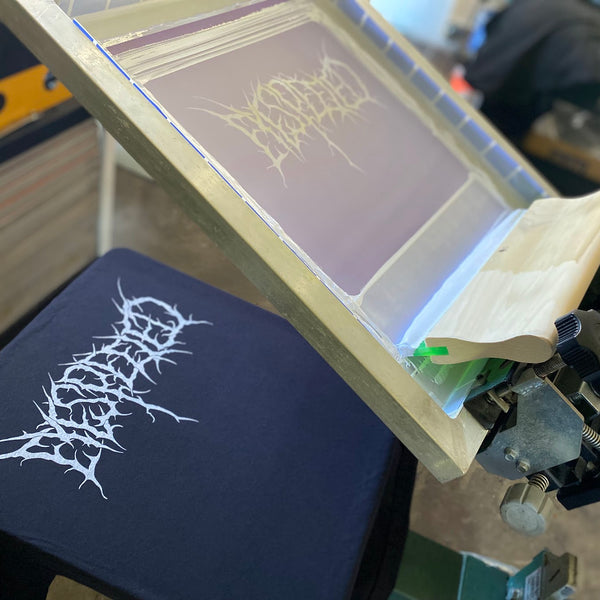 Metal band logo in white ink on a black tee on the press with the screen raised at RVA Threads screen printing, Virginia