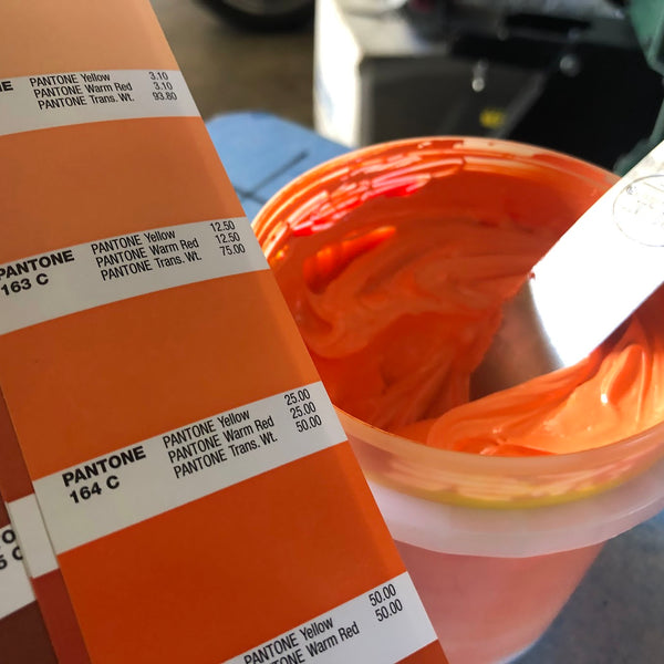 Orange ink mixed to match a Pantone swatch, with the spatula and swatch book at RVA Threads screen printing, Virginia