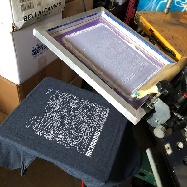 Richmond city line art in white ink on a navy tee with the screen raised on the press at RVA Threads screen printing, Virginia