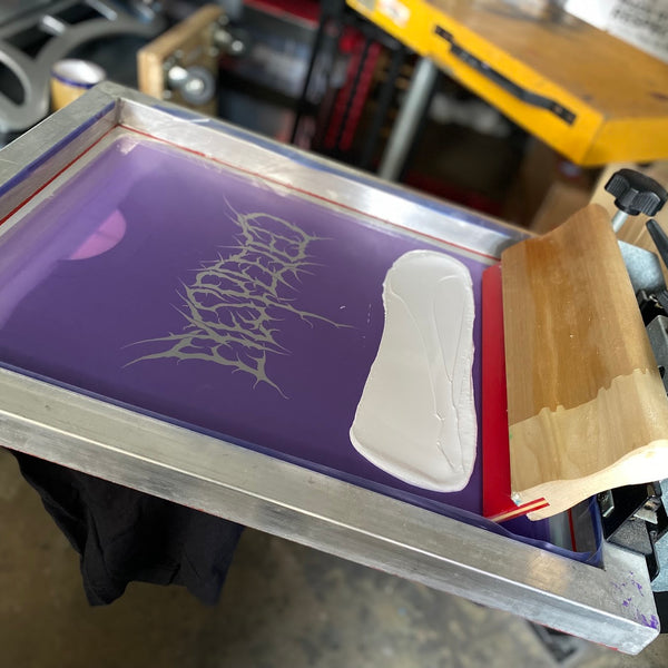 Screen with white ink and a squeegee on the press, gothic lettering design over black tees at RVA Threads screen printing, Virginia