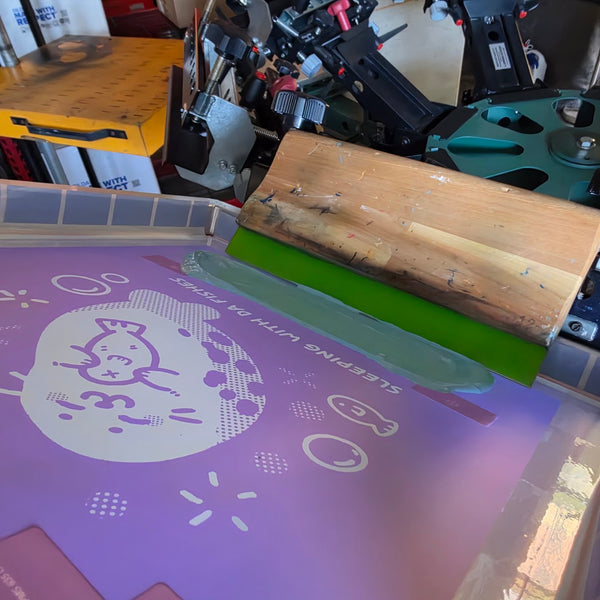 Sleeping With Da Fishes seal design screen with a green squeegee on the press at RVA Threads screen printing, Virginia
