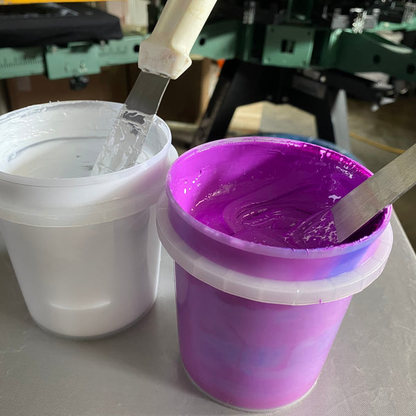 Tubs of white and purple ink with mixing sticks beside the press at RVA Threads screen printing, Virginia