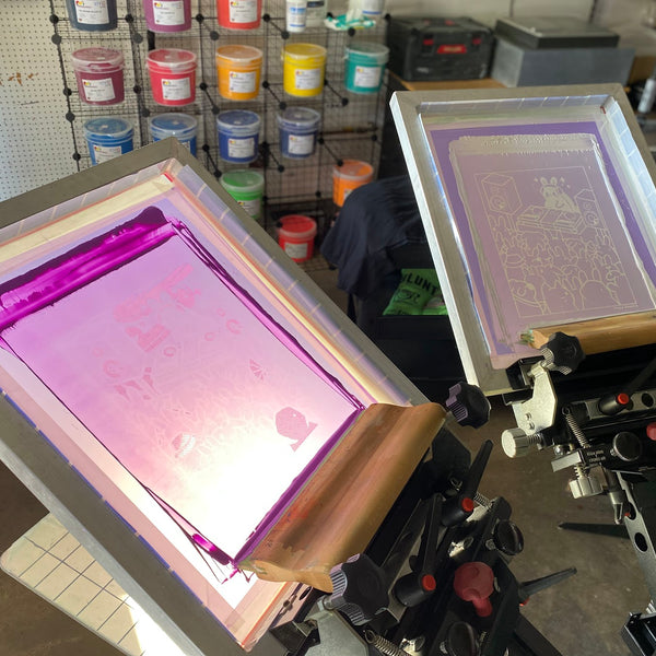 Two screens on the press, one flooded with pink ink, with the ink wall behind at RVA Threads screen printing, Virginia