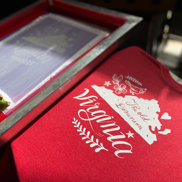 Virginia script design in white ink on a red tee beside the screen on the press at RVA Threads screen printing, Virginia