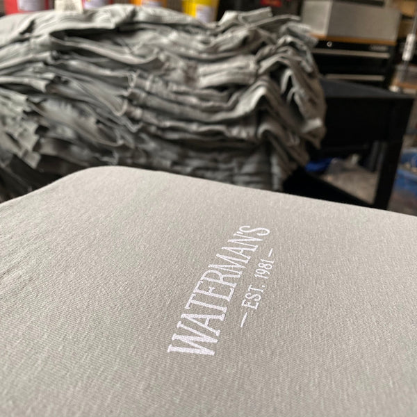 Waterman's left chest logo in white ink on a gray tee with the finished stack behind at RVA Threads screen printing, Virginia