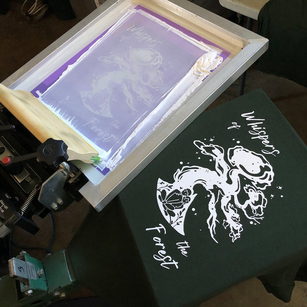 White ink illustration on a dark green tee with the screen raised on the press at RVA Threads screen printing, Virginia